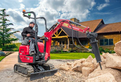 Red mini excavator with hydraulic arm operating on gravel near a rustic lodge building under blue sky