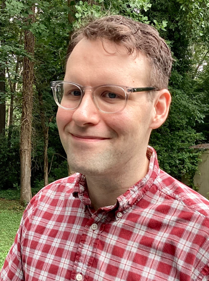 Man wearing glasses and red plaid shirt smiling outdoors with trees in background