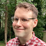 Man wearing glasses and red plaid shirt smiling outdoors with trees in background