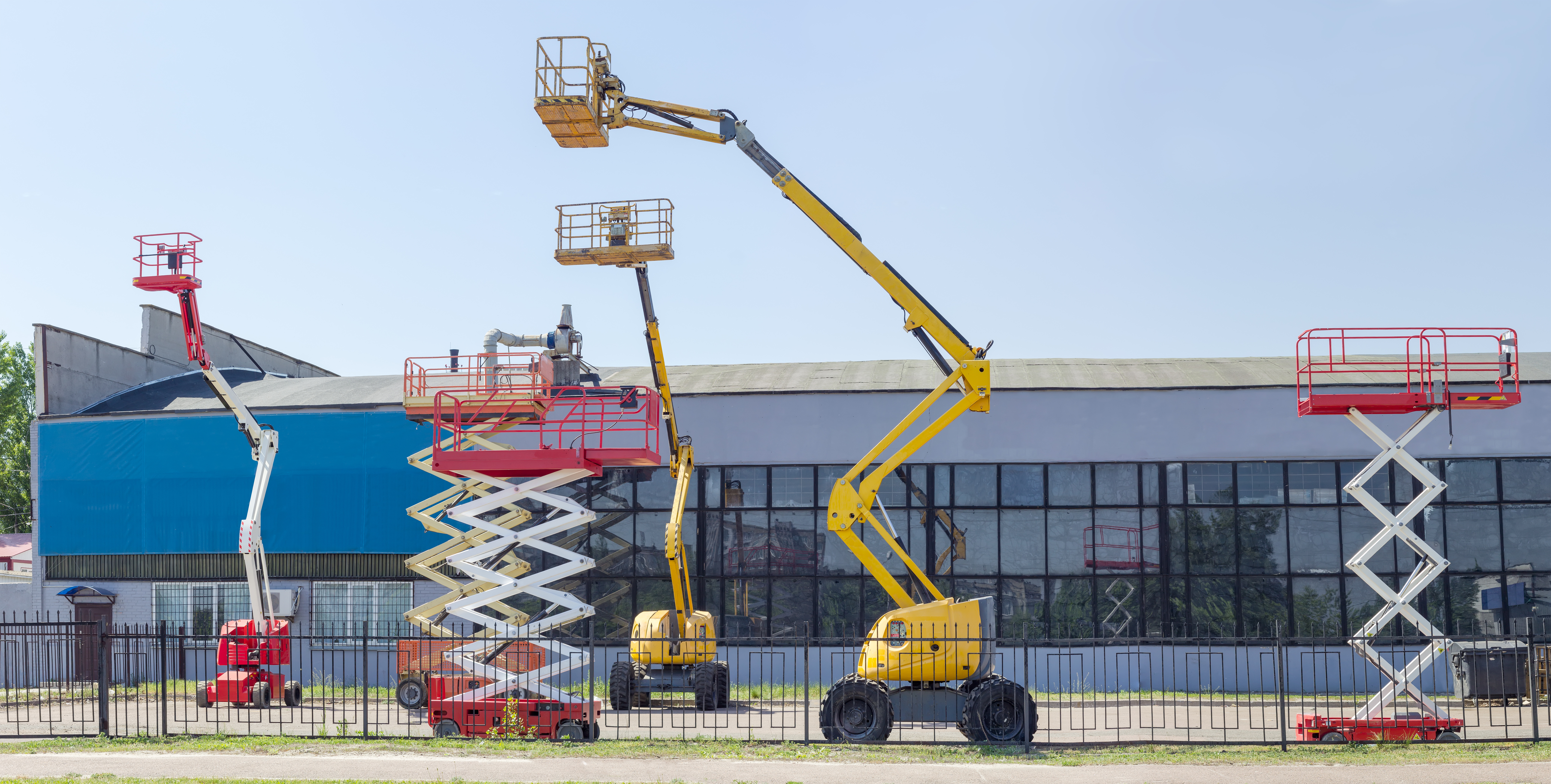 Construction equipment display yard featuring various aerial lifts and scissor lifts in red and yellow against industrial buildings.