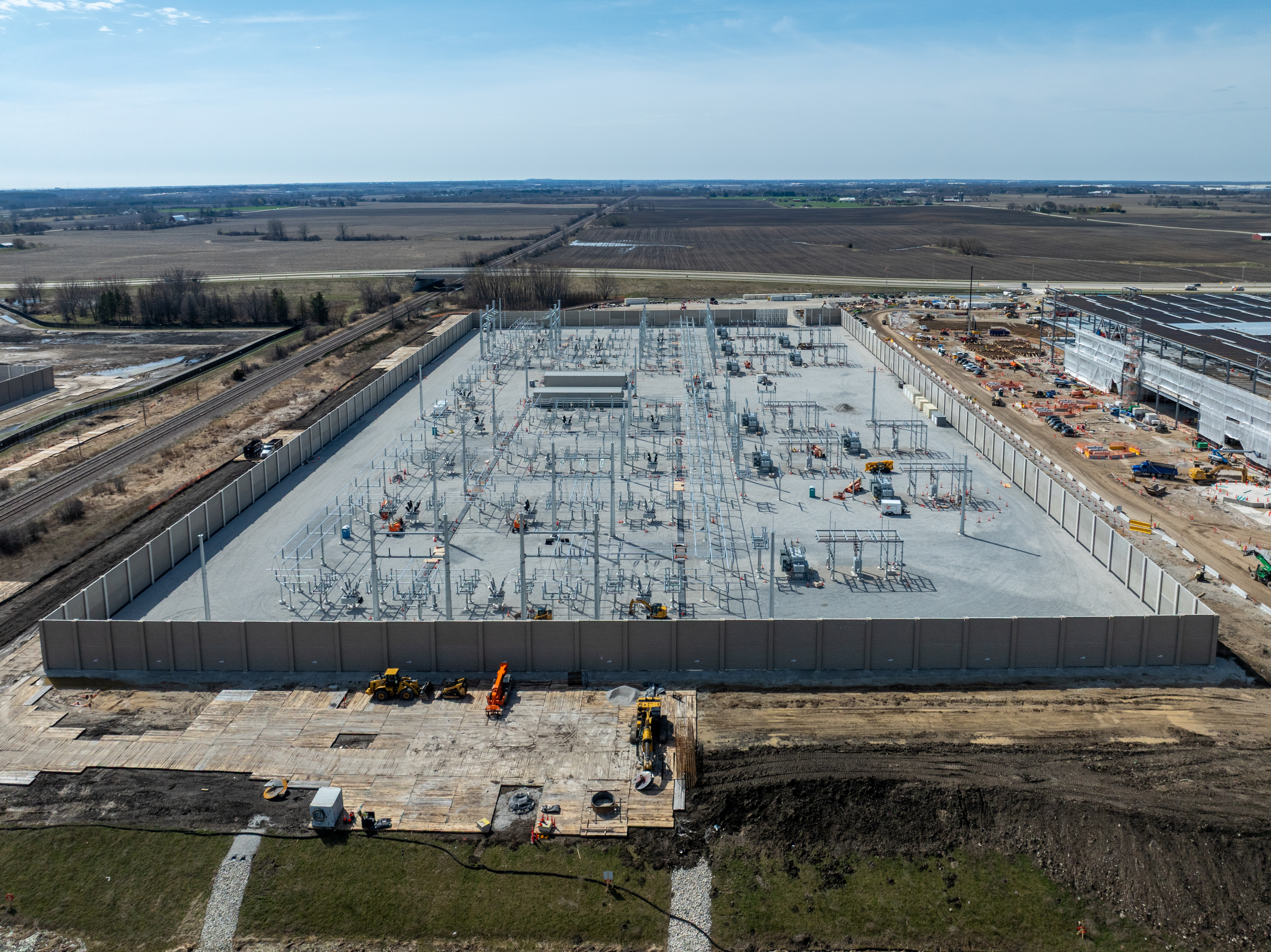 Aerial view of large industrial construction site with concrete foundation and electrical infrastructure surrounded by farmland