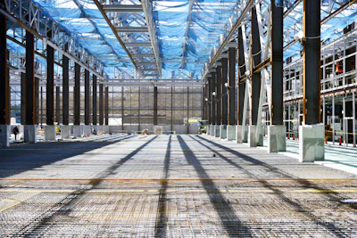 Interior of large industrial greenhouse with metal framework, blue plastic roof panels, concrete pillars, and paved flooring
