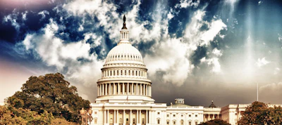 U.S. Capitol Building dome against dramatic cloudy sky with golden sunlight and surrounding trees