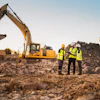 Construction site with two excavators, workers in safety vests, and dump trucks moving earth and rock materials