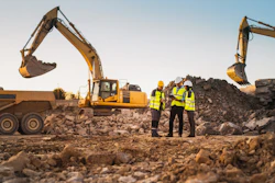 Construction site with two excavators, workers in safety vests, and dump trucks moving earth and rock materials