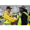 Two construction workers in safety gear shake hands during a workplace meeting in an industrial tent facility.