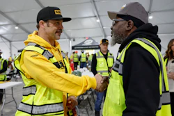 Two construction workers in safety gear shake hands during a workplace meeting in an industrial tent facility.