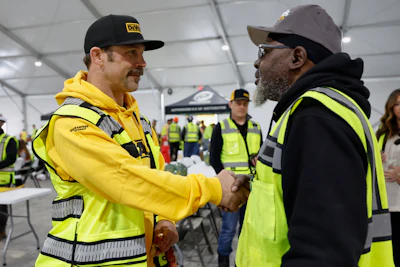 Two construction workers in safety gear shake hands during a workplace meeting in an industrial tent facility.