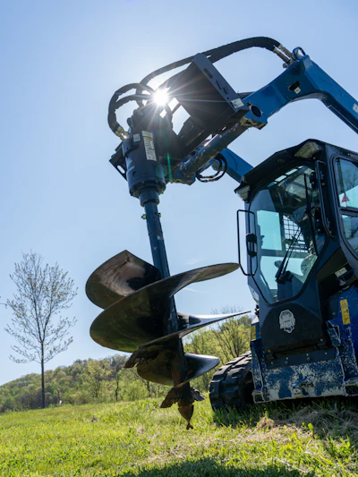Blue skid steer loader with auger drill attachment operating in a grassy field under clear sky