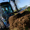 Blue skid steer loader moving pile of dark soil during landscaping work on hillside