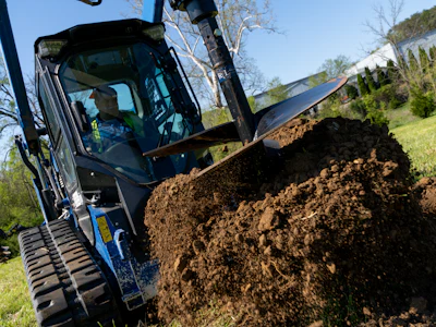 Blue skid steer loader moving pile of dark soil during landscaping work on hillside