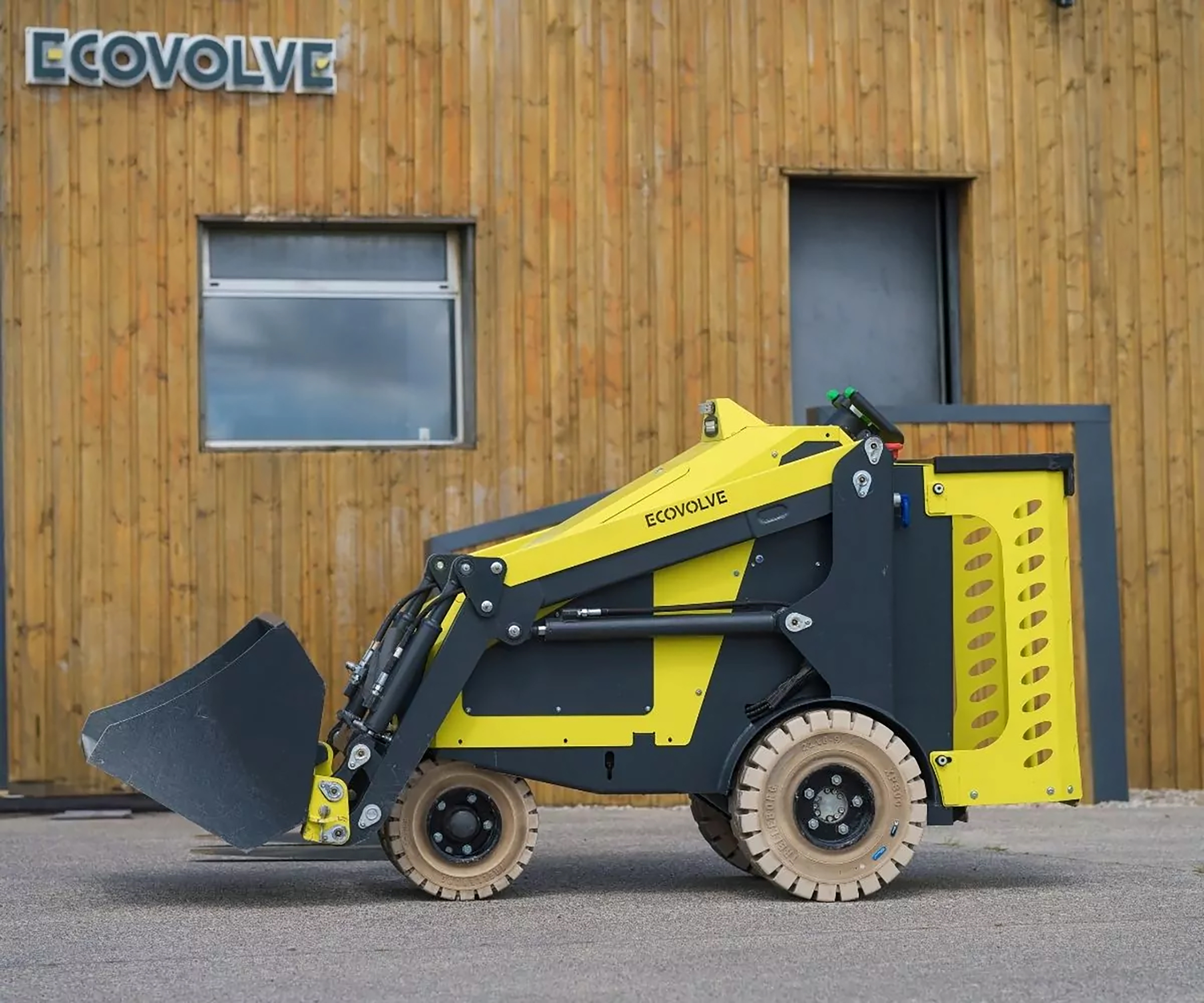 Yellow and black ECOVOLVE compact loader with bucket attachment positioned against wooden building