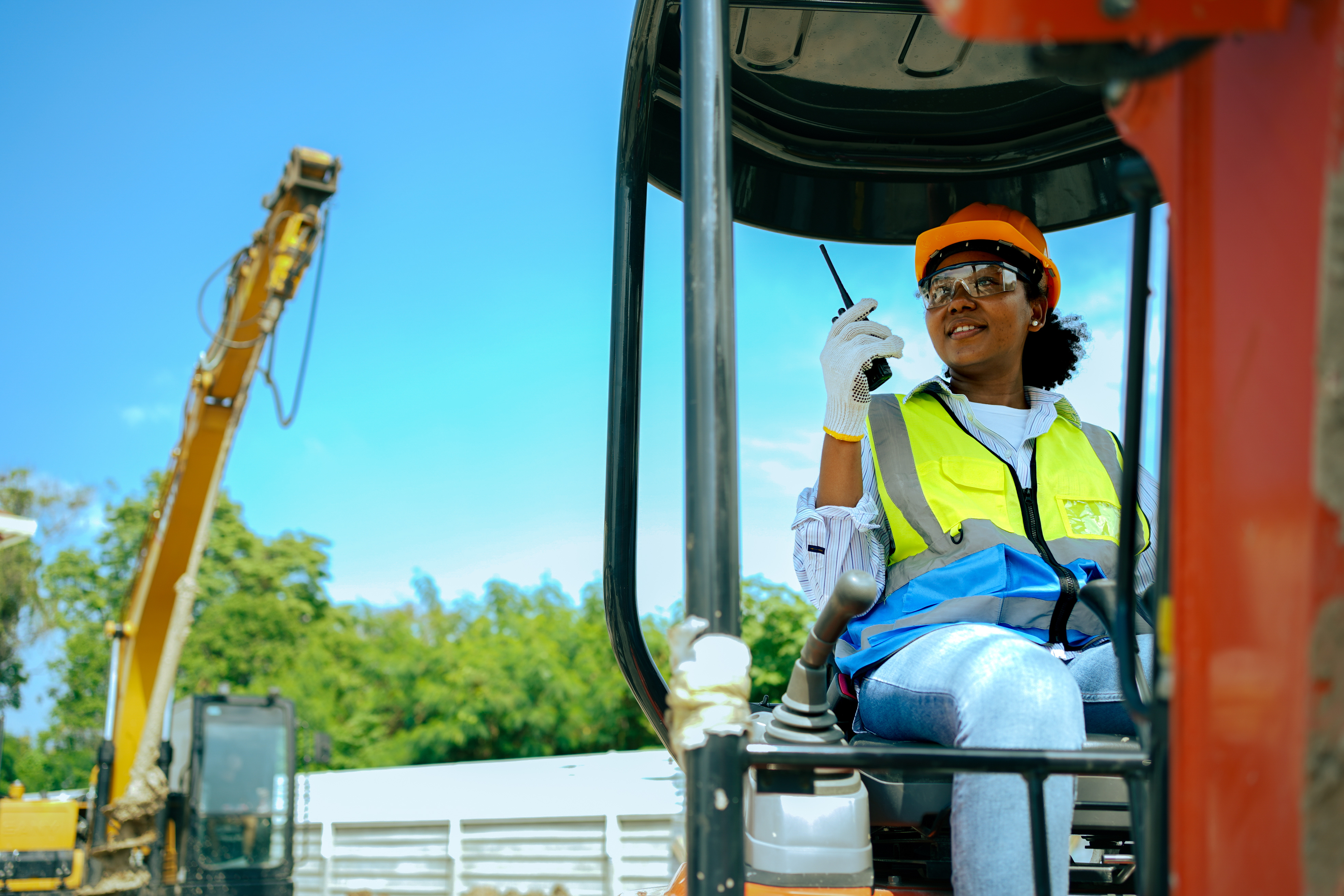 Construction worker in orange hard hat and safety vest operating heavy machinery from cabin with crane visible in background