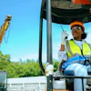 Construction worker in orange hard hat and safety vest operating heavy machinery from cabin with crane visible in background