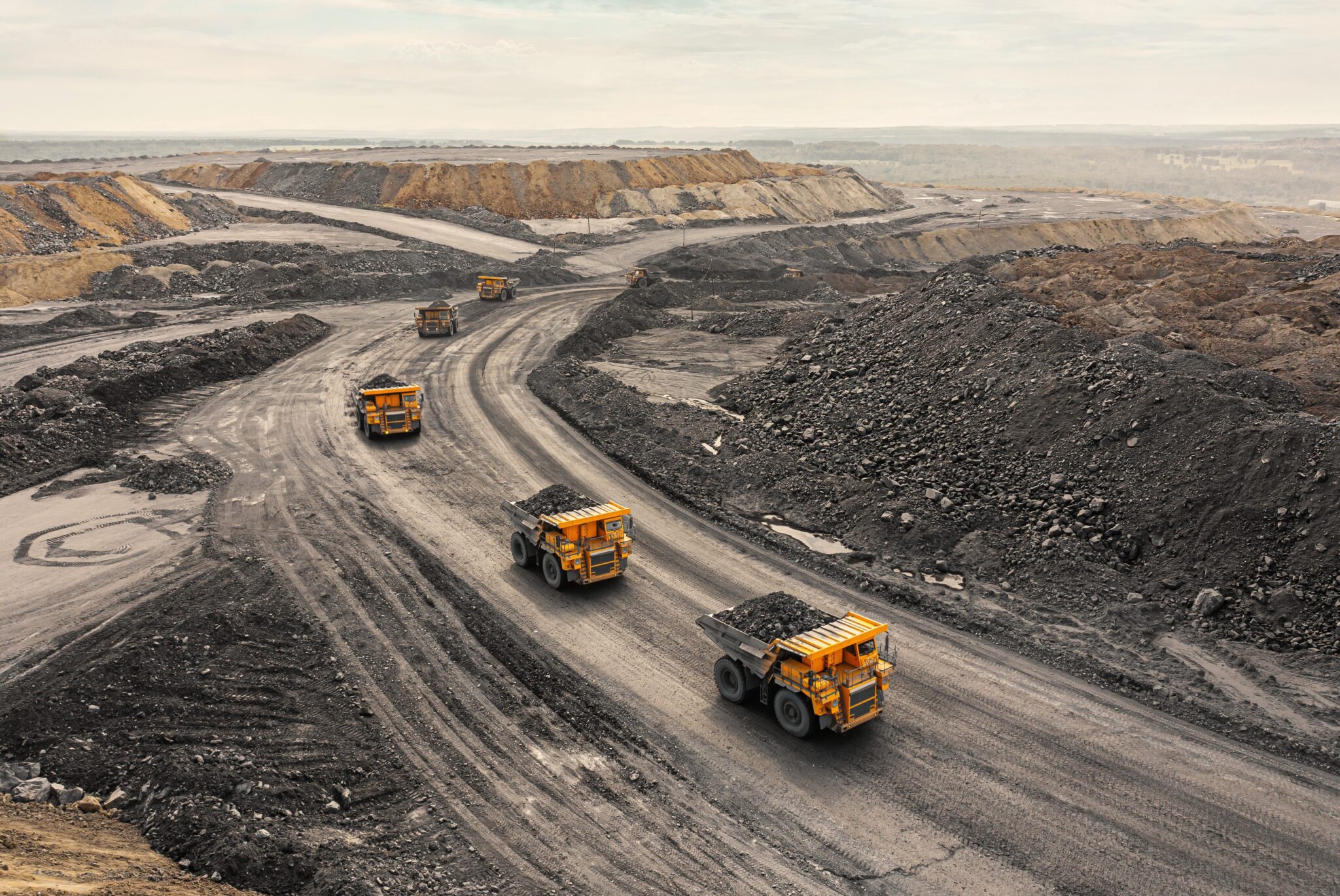 Heavy dump trucks navigate winding roads through an active quarry or mining site with excavated terrain and stockpiled materials.