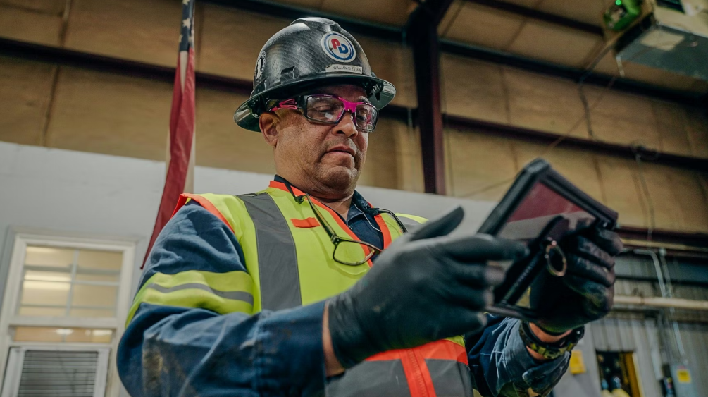 Industrial worker wearing safety helmet, glasses, and high-visibility vest holding equipment in warehouse