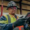 Industrial worker wearing safety helmet, glasses, and high-visibility vest holding equipment in warehouse
