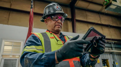 Industrial worker wearing safety helmet, glasses, and high-visibility vest holding equipment in warehouse
