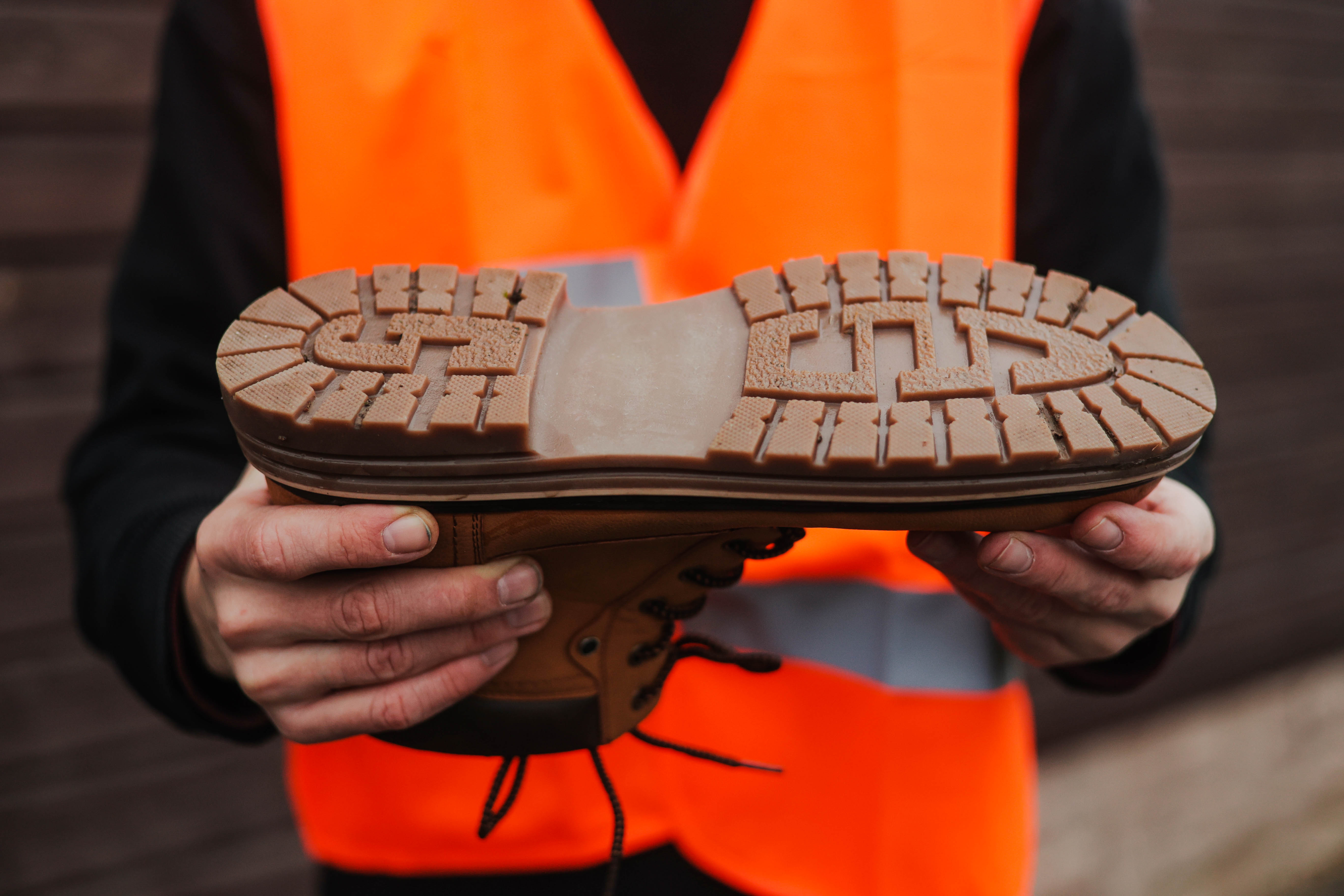 Worker in orange safety vest holding up brown shoe sole with tread pattern