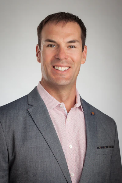 Professional headshot of a smiling man in gray blazer and pink shirt against neutral background