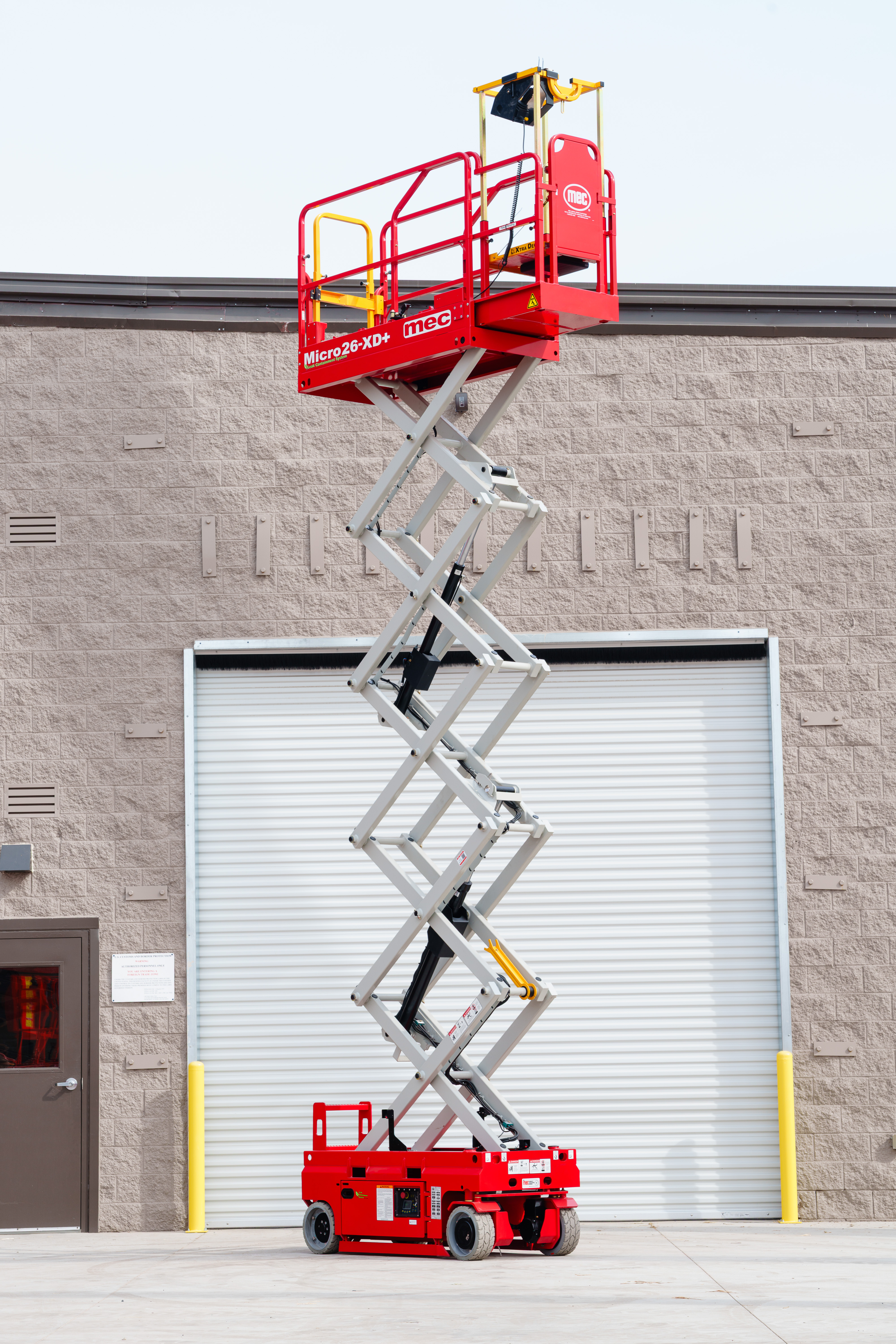 Red JLG Micro XD+ scissor lift fully extended against a brick building with white garage door