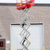 Red JLG Micro XD+ scissor lift fully extended against a brick building with white garage door
