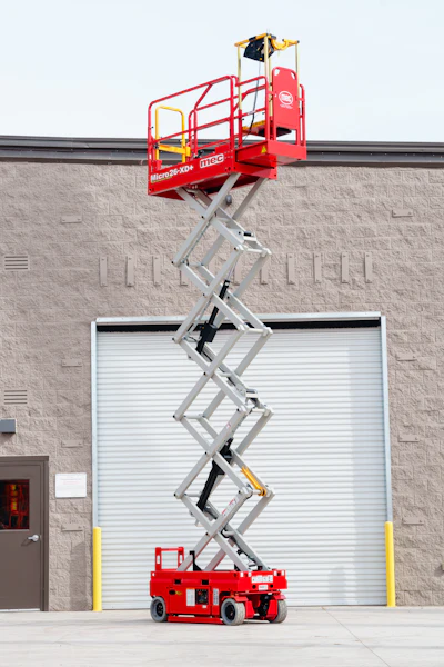 Red JLG Micro XD+ scissor lift fully extended against a brick building with white garage door