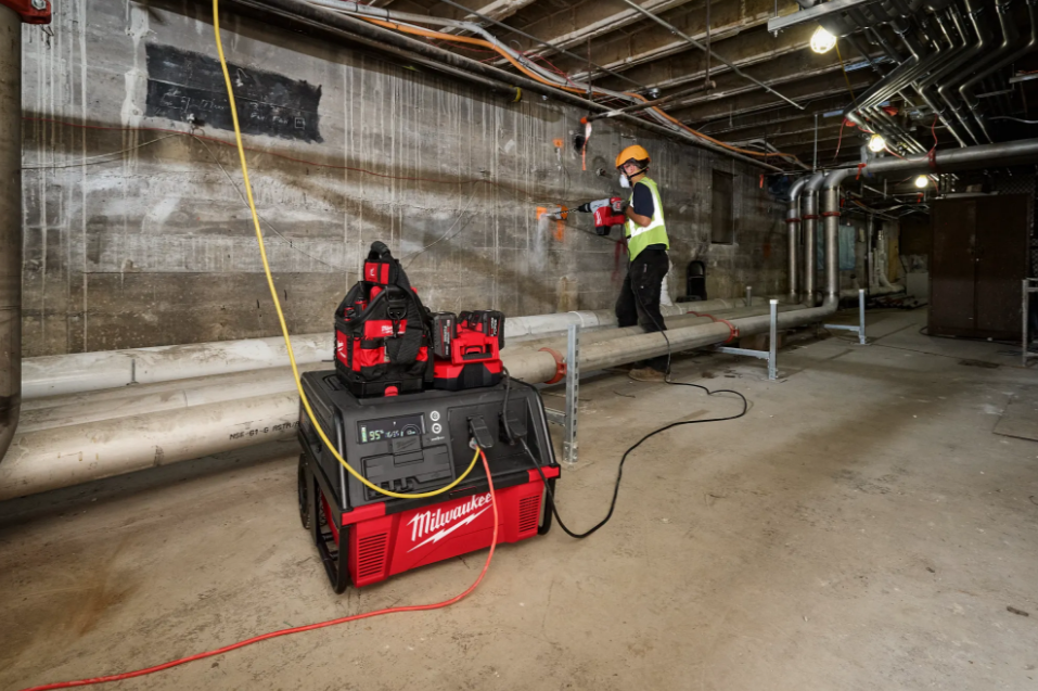 Construction workers using Milwaukee power equipment in an industrial building interior with concrete walls and exposed ceiling infrastructure.