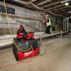 Construction workers using Milwaukee power equipment in an industrial building interior with concrete walls and exposed ceiling infrastructure.