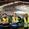 Four construction workers wearing safety vests and hard hats in an industrial warehouse discussing project plans
