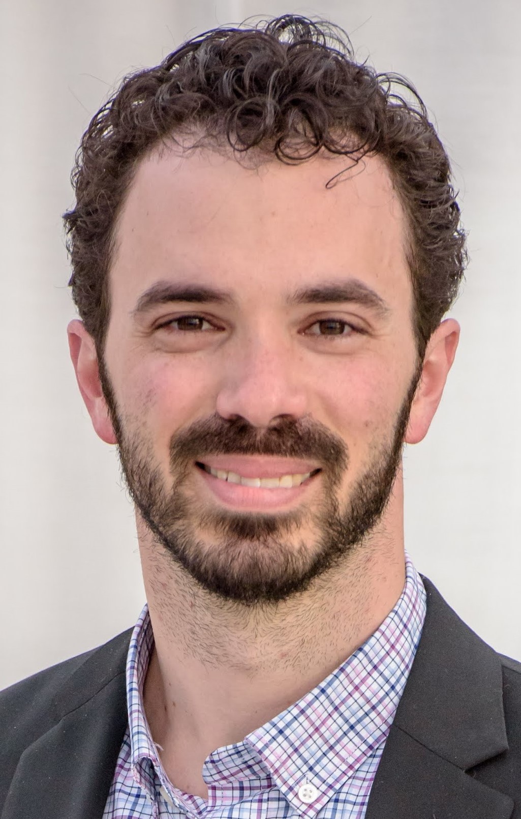 Professional headshot of a smiling man with curly dark hair and beard wearing a purple checkered shirt and dark blazer