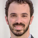 Professional headshot of a smiling man with curly dark hair and beard wearing a purple checkered shirt and dark blazer