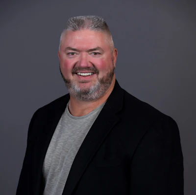 Professional headshot of a smiling man with gray hair and beard wearing a black blazer and gray shirt