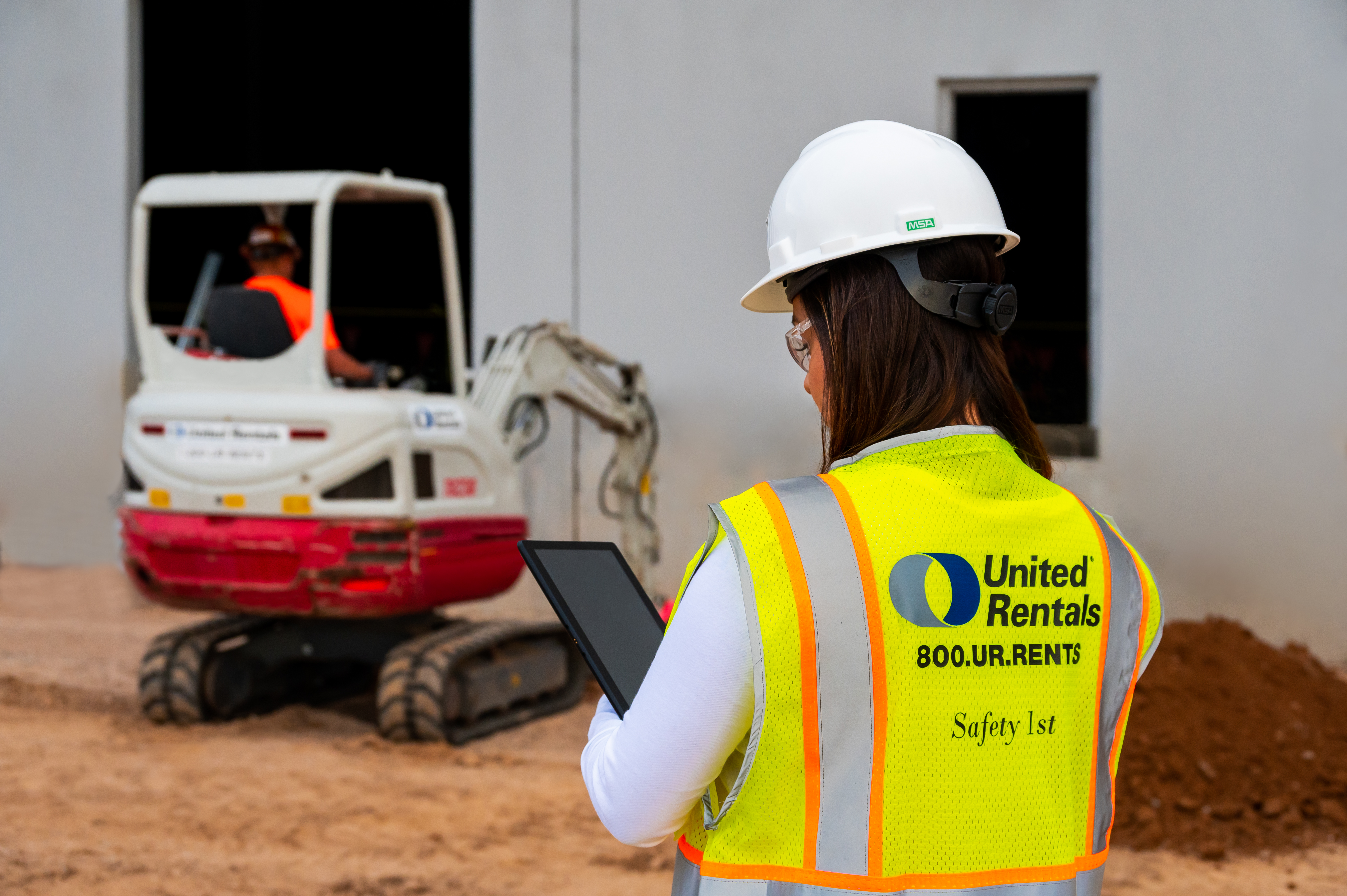 Construction worker wearing United Rentals safety vest and hard hat operating excavator on job site