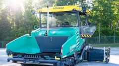 Turquoise VÖGELE asphalt paver with yellow crane attachment parked outdoors near trees