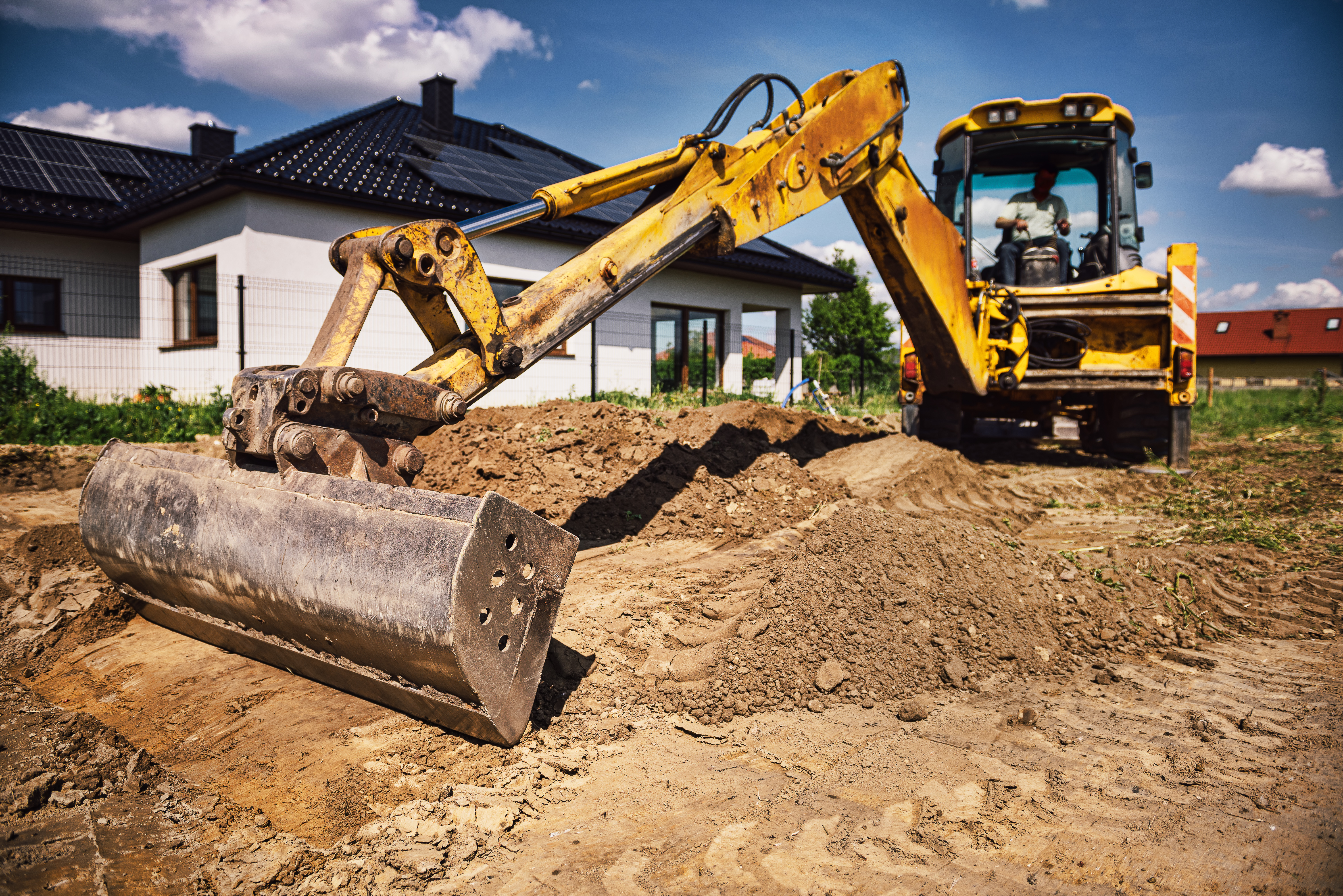 Yellow excavator with compactor attachment working on residential construction site with white house in background