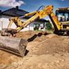 Yellow excavator with compactor attachment working on residential construction site with white house in background