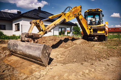 Yellow excavator with compactor attachment working on residential construction site with white house in background