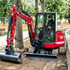 Red compact excavator with operator working on tree removal in a park surrounded by green trees and orange safety fencing