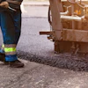 Asphalt paving machine laying road surface with dust clouds and construction worker in safety vest