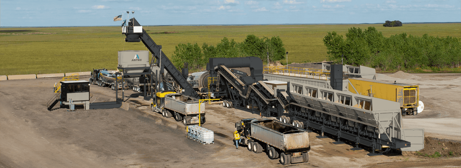 Industrial asphalt paving facility with mixing equipment, conveyor systems, and trucks on a construction site.