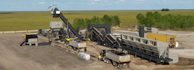 Industrial asphalt paving facility with mixing equipment, conveyor systems, and trucks on a construction site.