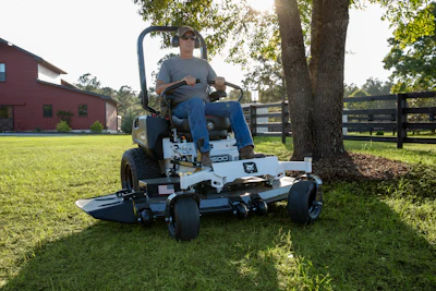 Man operating a zero-turn radius mower on a grassy farm with red barn and wooden fence in background