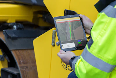 Worker in safety vest holding surveying tablet with GPS mapping software near yellow equipment