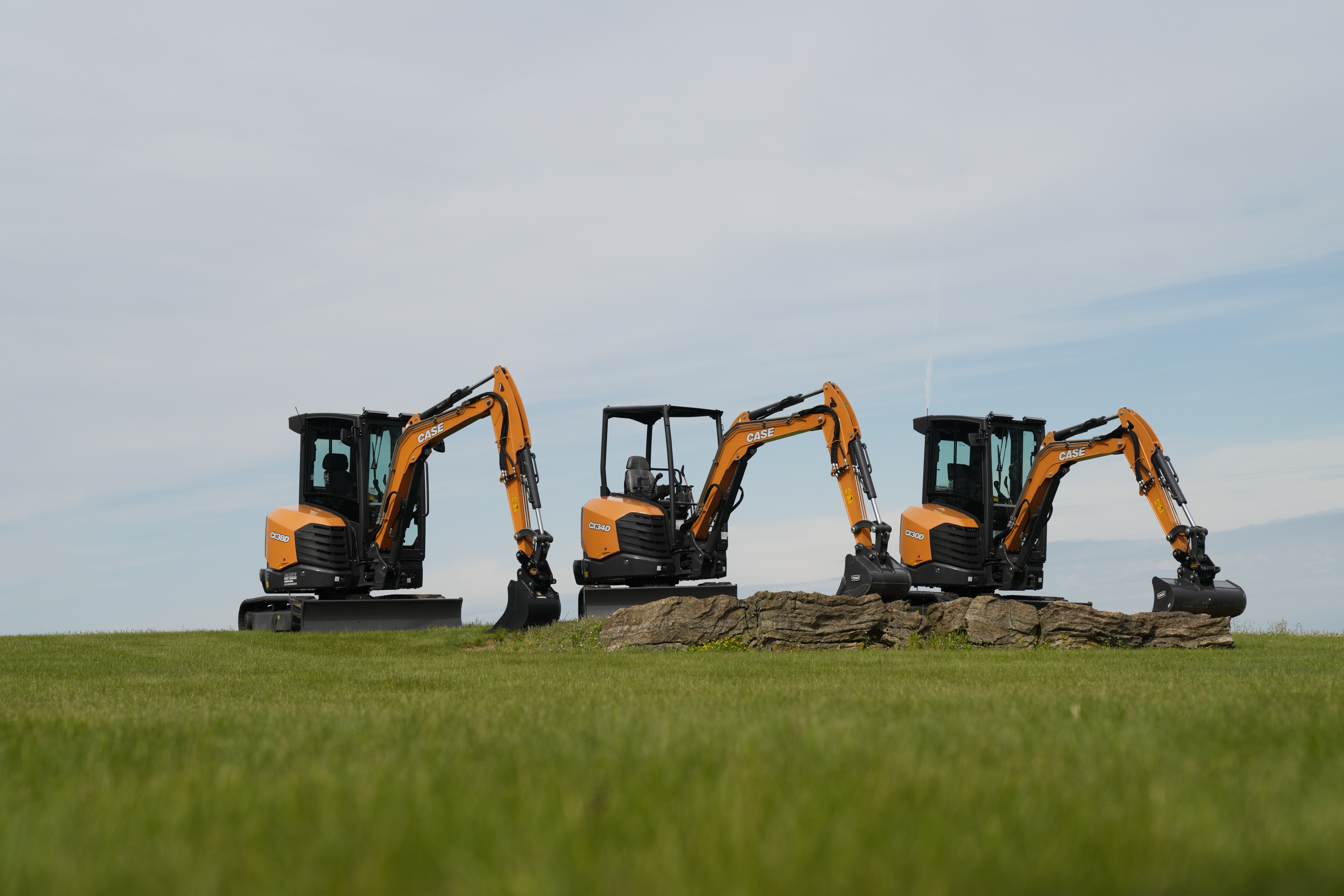 Three orange and black compact excavators lined up in a green field under a partly cloudy sky