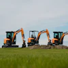 Three orange and black compact excavators lined up in a green field under a partly cloudy sky