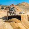 Bulldozer moving gravel at a quarry with piles of earth and equipment visible on a sunny day