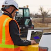Construction worker wearing safety helmet and orange vest using laptop at outdoor job site with excavator in background