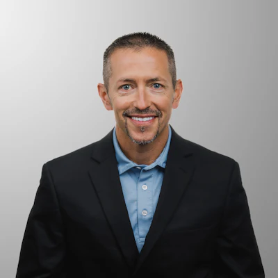Professional headshot of a smiling man in black blazer and light blue shirt against neutral background