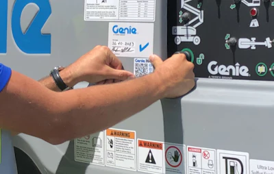 Person pointing to QR code and Genie label on industrial lift equipment with warning signs